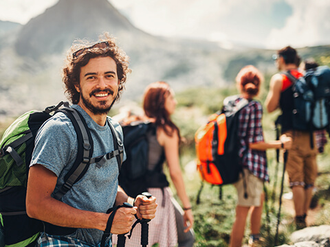 Ein Mann mit längerem Haar lächelt in die Kamera, während er mit seinen Freunden auf einen Berg hinauf wandert.
