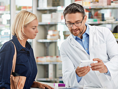 Eine junge blonde Dame wird in der Apotheke von einem Apotheker mit Brille beraten. Sie schauen sich gemeinsam ein Medikament an.