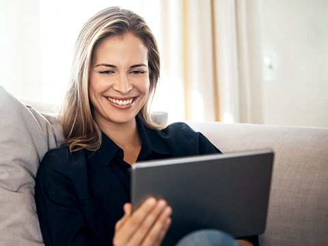 Eine Frau sitzt auf der Couch und sieht sich etwas auf einem Tablet-Computer an. Sie lächelt dabei.