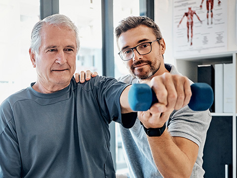 Ein Physiotherapeut leitet einen älteren Mann beim Krafttraining an. Er streckt den Arm aus und hebt einen Hantel.